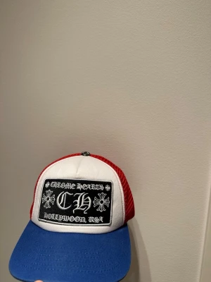 Chrome Hearts truckerkeps röd/blå/vit - Snygg truckerkeps från Chrome Hearts med vit front, blå skärm och röda meshpaneler. Stor svart patch framtill med Chrome Hearts-logga och blommotiv samt texten 'Hollywood, USA'. Justerbar baktill för perfekt passform.