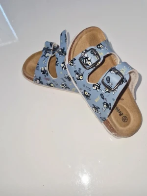Blå sandaler med Badtz-Maru motiv - Säljer ett par blå sandaler med dubbla spännen och gulligt Badtz-Maru mönster. Sandalerna har en bekväm fotbädd i mockaimitation och justerbara remmar. Perfekta för dig som gillar unika och lekfulla detaljer på dina skor.