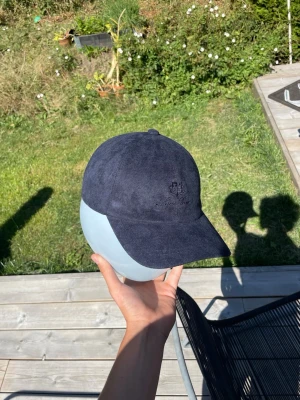 Mörkblå mockakeps med broderi - Snygg mörkblå keps i mockaimitation med böjd skärm och diskret broderad logga framtill. Klassisk sexpaneldesign och justerbar rem bak. Perfekt accessoar för en clean och stilren look.