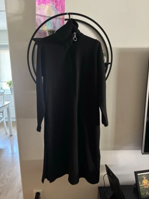 Svart långärmad ribbad klänning - Svart långklänning med ribbad struktur och hög krage. Klänningen har långa ärmar och en dekorativ dragkedja vid halsen. Perfekt för dig som gillar en stilren och enkel look. I fint skick! 