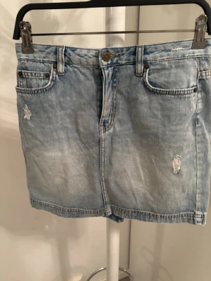 Ljusblå jeanskjol med slitningar - Säljer en ljusblå jeanskjol i kort modell med slitna detaljer framtill. Kjolen har klassiska fem fickor, knapp och dragkedja i midjan. Perfekt för dig som gillar en avslappnad och trendig stil.