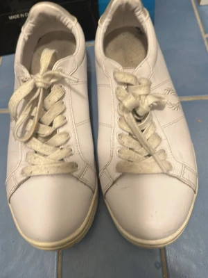 Vita Fred Perry sneakers i läder - Klassiska vita sneakers från Fred Perry i läder med den ikoniska lagerkransen broderad på sidan. Skorna har vita snören, låg profil och diskreta sömmar för en clean look. Perfekta till jeans eller shorts för en stilren vibe.