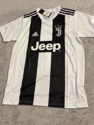 Juventus matchtröja Ronaldo 7 Adidas - Juventus officiella matchtröja i vitt och svart med breda ränder, Adidas-logga och Jeep som sponsor. På ryggen står det 'Ronaldo' och nummer 7. Tillverkad i lätt och ventilerande material, perfekt för fotbollsträning eller match.