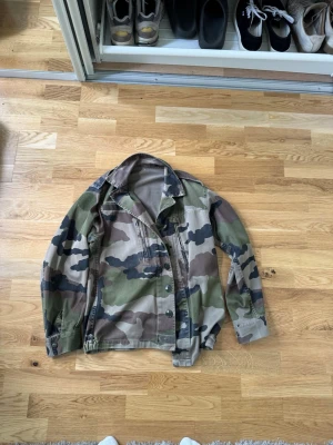 Camouflage overshirt med knappar - Säljer en overshirt i klassiskt camouflage-mönster med gröna, bruna och beige toner. Skjortan har krage, knappstängning framtill och långa ärmar. Perfekt för en streetwear-look eller lager på lager.