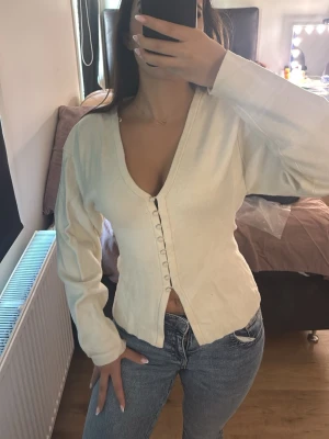 Vit cardigan med knappar - Stilren vit cardigan med djup v-ringning och dekorativa knappar framtill. Långärmad modell i mjukt material som sitter snyggt på kroppen. Perfekt att matcha med jeans eller kjol för en clean look.