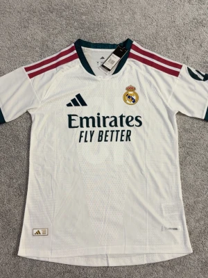 Real Madrid matchtröja vit Adidas - Säljer en officiell Real Madrid matchtröja från Adidas i vitt med mörkgröna detaljer och vinröda ränder på axlarna. Tröjan har klubbmärke på bröstet, sponsortryck och är tillverkad i ett lätt, ventilerande funktionsmaterial.