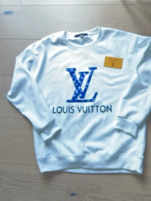 Vit sweatshirt från Louis Vuitton - Säljer en vit sweatshirt från Louis Vuitton med stort blått LV-tryck och logga framtill. Tröjan har rund halsringning och långa ärmar. Materialet ser ut att vara mjukt och bekvämt, perfekt för en trendig och avslappnad stil. Och lite smuts fläckar 