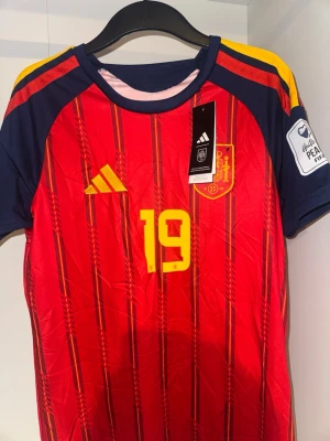 Spansk landslagströja Lamine Yamal - Röd och marinblå matchtröja från Adidas för Spaniens landslag, med gula detaljer och nummer 19. Lamine Yamal står tryckt på ryggen. Tröjan har korta ärmar, officiell landslagssköld och FIFA-märke på ärmen. 