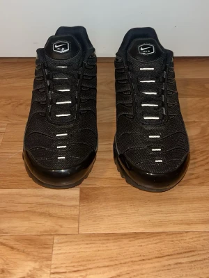Nike Air Max Plus - Säljer ett par svarta Nike Air Max Plus sneakers med mesh- och syntetmaterial, ikoniska vågiga detaljer och synlig Air-dämpning i sulan. Skorna har vita detaljer på snörningen och en chunky sula som ger en sportig vibe.