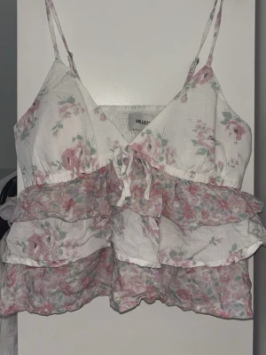 Blommig volangtop från Hollister - Söt blommig topp från Hollister med tunna axelband och volanger. Perfekt för sommaren och ger en luftig känsla tack vare det tunna materialet. 🎀