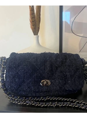 Svart axelväska i bouclé - Säljer en svart axelväska i bouclétyg med silverfärgad kedjerem och vridlås framtill. Väskan har en mjuk, fluffig textur och en klassisk rektangulär form. Perfekt för dig som gillar trendiga accessoarer med lite extra attityd.