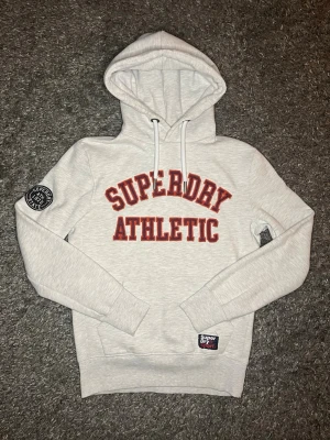 Ljusgrå hoodie från Superdry - Ljusgrå hoodie från Superdry med röd text 'Superdry Athletic' på bröstet och patch på ärmen. Klassisk huva med vita dragsnören och ribbade muddar. Mjuk bomullsblandning och Superdry-märke vid nederkanten. Perfekt för en avslappnad stil.