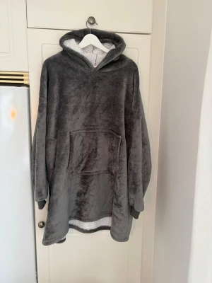 Två st mysiga grå oversized hoodie-filtar - Supermjuk och fluffig grå hoodie-filt med stor känguruficka framtill och huva. Perfekt för att chilla hemma eller mysa i soffan. Insidan är fodrad med vit teddy för extra värme och komfort. Långärmad och oversized modell för maximal mysfaktor. Säljes för 200 kr styck.