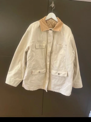 Arket canvasjacka med manchesterkrage - Stilren beige canvasjacka med ljusbrun manchesterkrage och flera praktiska fickor framtill. Jackan har knappar och lång ärm, perfekt för lager på lager under vår och höst. Tillverkad i bomull för en avslappnad och bekväm look. Några små fläckar (se bilder) men dessa syns ej när den är på. Jackan är nytvättad så fläckarna har inte gått bort i tvätt, men kanske att kemtvätt kan ta bort dem. 
