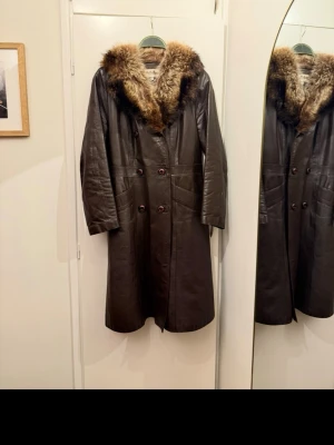 Brun skinnkappa med päls - Retro snygg brun äkta skinnkappa från Terhi Riini med stor, fluffig äkta päls runt kragen. Kappan har dubbelknäppning framtill och två fickor. Perfekt för kalla dagar när du vill ha både stil och värme. Storlek 38 men liten i storlek mer som en 36a