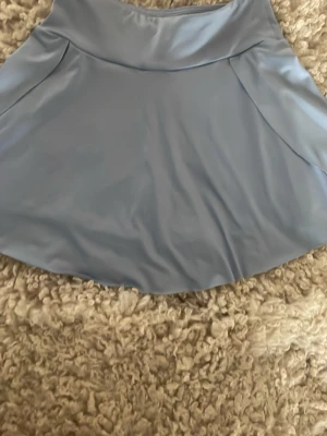 Ljusblå skort med plisserad baksida - Supersnygg ljusblå skort med slät framsida och plisserad baksida. Kjolshortsen har en hög midja och är gjorda i ett glansigt, stretchigt material som ger en sportig vibe. Perfekt för dig som gillar en trendig och bekväm stil. Helt ny aldrig använd.