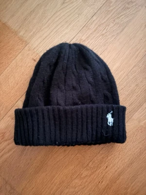  Polo Ralph Lauren mössa  - Svart kabelstickad mössa från Polo Ralph Lauren med uppvikt kant och broderad vit logga framtill. Klassisk och stilren design som passar perfekt till kalla dagar. Tillverkad i mjukt material för extra komfort.