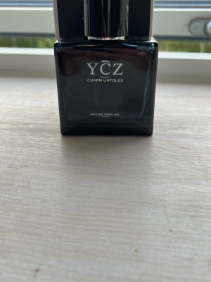 YCZ Charm Unfolds parfym - Snygg parfymflaska från YCZ med texten 'Charm Unfolds'. Flaskan är mörkblå och har en stilren, fyrkantig form med svart och silverfärgat lock. Doften heter Marine Perfume och ger en fräsch känsla. Perfekt för dig som gillar exklusiva och moderna dofter.
