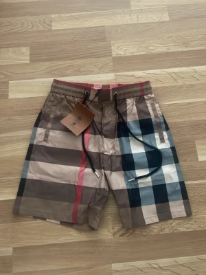 Rutiga burberry shorts med snörning - Snygga shorts med stort rutmönster i beige, svart, brun, vit och en röd detalj. Shortsen har elastisk midja med snörning och sidofickor. Perfekta för sommaren och ger en trendig look.