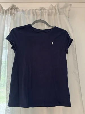 Mörkblå t-shirt från Ralph Lauren - En stilren mörkblå t-shirt från Ralph Lauren med klassisk passform och korta ärmar. T-shirten har en liten vit broderad logga på bröstet och är gjord i mjuk bomull. Jätte fint skick. Skulle säga att det passar xxs/xs också