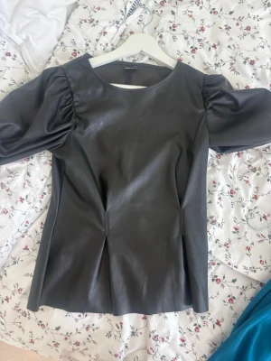 Svart puffärmad topp Gina Tricot - Säljer en svart topp från Gina Tricot i skinnimitation med puffärmar och rund hals. Toppen har en figurnära passform och är perfekt för dig som vill ha en edgy look. Materialet är syntet och ger en cool känsla till outfiten.