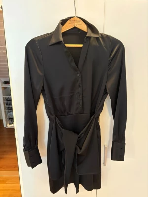 Svart omlottklänning med knyt - Stilren svart kortklänning i satinliknande material med lång ärm, krage och knappar framtill. Klänningen har ett snyggt knyt i midjan som ger en markerad siluett och en elegant look. Perfekt för dig som gillar minimalistisk och chic stil.