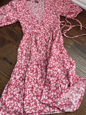 Rosa blommig klänning från Gina Tricot - Supersöt rosa omlottklänning från Gina Tricot med vitt blommönster. Klänningen har v-ringning, knytband i midjan och korta puffärmar. Perfekt för varma dagar och ger en romantisk vibe till din outfit.