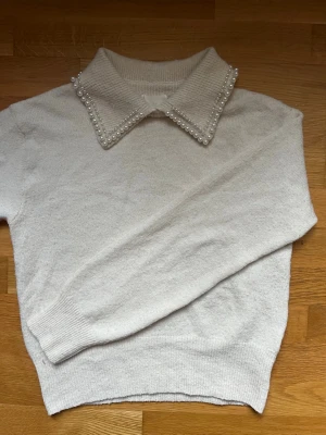 Vit stickad tröja med pärlkrage H&M - Supersöt vit stickad tröja från H&M med en unik krage dekorerad med pärlor. Tröjan har långa ärmar, ribbade muddar och är gjord i ett mjukt material som känns skönt mot huden. Perfekt för dig som vill ha något extra till din outfit.