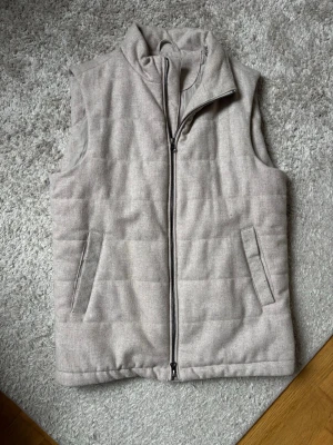 Beige dunväst från John Henric - Stilren dunväst från John Henric med hög krage och två snedställda fickor framtill. Västen har en mjuk yta, quiltad design och dragkedja hela vägen. Perfekt för lager på lager och enkel att matcha med olika outfits.