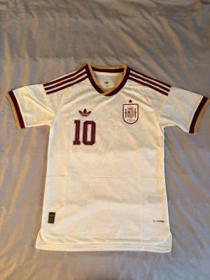 Lamine yamal - Vit Spanien tröja nr 10 Adidas - ”feltryckt” - Säljer en vit Spanien fotbollströja från Adidas med vinröda detaljer, nummer 10 och Lamine Yamal på ryggen. Tröjan har korta ärmar, ribbade muddar och klassiska Adidas-ränder på axlarna. Perfekt för dig som älskar fotboll och vill sticka ut på planen.
