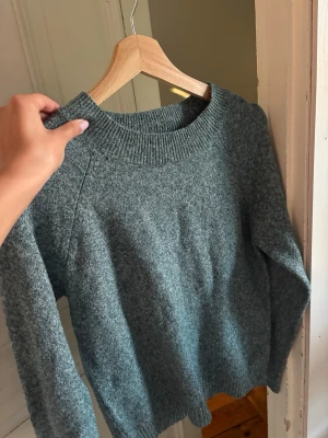 Grönmelerad stickad tröja - Mysig grönmelerad stickad tröja med rund halsringning och ribbade muddar vid ärmslut och nederkant. Tröjan har en avslappnad passform och är perfekt för kyligare dagar. Materialet ser ut att vara en mjuk ull- eller ullblandning.