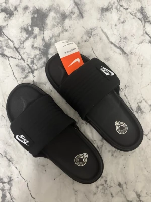 Nike tofflor - Säljer ett par svarta sandaler från Nike med bred, vadderad rem och vit logga på ovansidan. Sandalerna har en klassisk slip-in design och är tillverkade i syntetmaterial med bekväm innersula. Perfekta för sommaren eller att slappa i. Helt nya med tags på, storlek 43