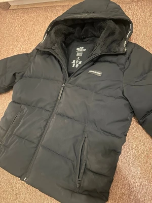 Vinterjacka  - Säljer en svart pufferjacka från Hollister med mjukt foder och huva. Jackan har dragkedja framtill, två sidofickor med dragkedja och Hollister-logga på bröstet. Perfekt för kalla dagar och riktigt skön att bära.