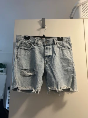Ljusblå denimshorts med slitningar - Säljer ett par ljusblå denimshorts med råa kanter och slitningar framtill och baktill. Shortsen har fem fickor, bälteshällor och knappgylf. Perfekta för sommaren och ger en avslappnad vibe.