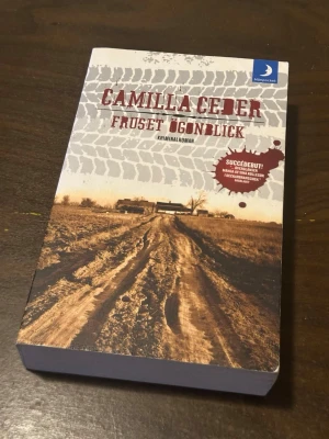 Fruset ögonblick av Camilla Ceder - En kriminalroman i pocketformat av Camilla Ceder, utgiven av Månpocket. Omslaget har en brun och beige ton med ett lantligt motiv och tydliga däckspår i leran. Boken handlar om ett mordfall på landsbygden och är Ceders debutroman.