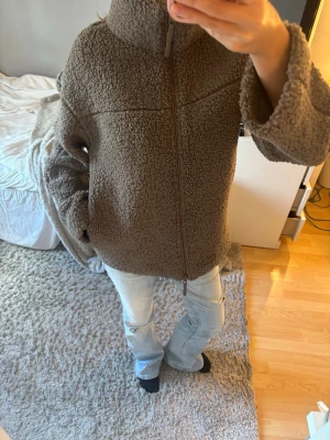 Brun teddyjacka med dragkedja - Mysig brun teddyjacka med hög krage och hel dragkedja framtill. Jackan har en oversized passform och är gjord i fluffigt teddy-material som ger en avslappnad och trendig look. Perfekt för kalla dagar när du vill vara både varm och snygg.