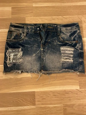 Blå jeanskjol med slitningar - Säljer en kort blå jeanskjol med slitna detaljer och dekorativa nitar på ena sidan. Kjolen har dragkedja, knappen har ramlat av men går lätta tt sy fast en ny knapp om man vill. Köpt utomlands. 
