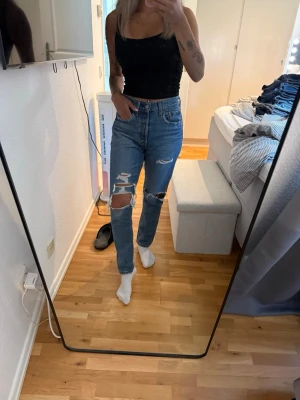 Blå jeans med slitningar - Säljer ett par ljusblå mom jeans med hög midja och raka ben. Jeansen har flera slitningar och hål på båda benen, vilket ger en trendig och avslappnad look. Klassisk femficksmodell och stängs med dragkedja och knapp. Modell 501 i märket Levis W27L28