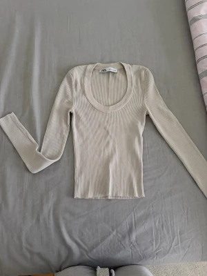 Beige ribbad topp från Zara - Säljer en beige ribbad långärmad topp från Zara. Toppen har en djup rundringning och är tillverkad i ett mjukt, stretchigt material som sitter snyggt på kroppen. Perfekt att styla med jeans eller kjol för en clean look. Aldrig använd