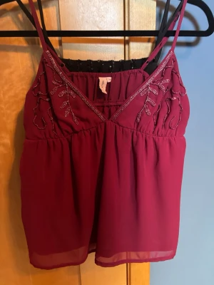 Vinröd linnetopp med pärldetaljer - Snygg vinröd linnetopp med tunna axelband och v-ringning. Toppen har pärldetaljer och broderade mönster upptill som ger en unik look. Tillverkad i ett lätt och luftigt material, perfekt för varma dagar eller lager på lager.