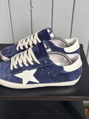 Blåa sneakers med vit stjärna - Snygga blåa sneakers från Golden Goose med vit stjärndetalj på sidan och vita skosnören. Skorna har en låg siluett och är tillverkade i mocka med kontrasterande vita detaljer på häl och sula. Perfekta för en trendig och avslappnad look.