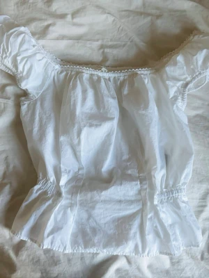 Vit offshoulder blus med resår - Söt vit offshoulder blus med resår i midjan och vid ärmsluten. Blusen har en sommar känsla med små spetsdetaljer längs urringningen och är perfekt för varma dagar. Tillverkad i ett lätt bomullstyg som känns luftigt och bekväm.