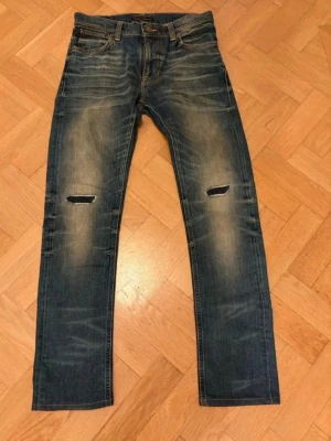 Blå slitna jeans med hål på knäna - Säljer ett par blå jeans med slitningar och hål på båda knäna. Jeansen har en smal passform, klassisk femficksmodell och tydliga slitningar även på bakfickorna. Perfekta för en avslappnad och trendig look.