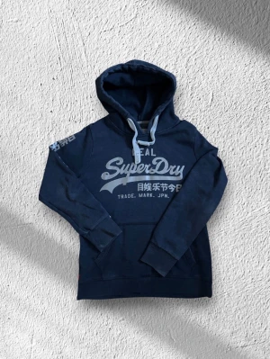 Mörkblå hoodie från Superdry - Snygg mörkblå hoodie från Superdry med stor logga och japansk text på bröstet. Hoodien har vita snören i huvan och en klassisk känguruficka framtill. Perfekt för en avslappnad och cool stil.