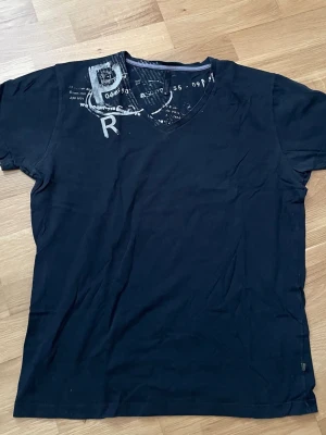 Svart t-shirt med tryck och v-ringning, XL Jack&Jones  - Säljer en svart/ mörkblå  t-shirt med v-ringning och coolt tryck i vitt och grått vid axeln. T-shirten har korta ärmar och är gjord i mjuk bomull, perfekt för en avslappnad stil.