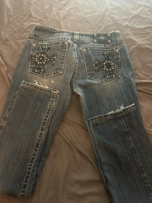 Blå miss me jeans med korsdetaljer bak - Unika blå jeans med slitna detaljer och stora kors i silvernitar på bakfickorna. Jeansen har en rak passform.. Perfekta för dig som vill sticka ut med coola detaljer och streetwear-vibe. Dem sitter jättebra