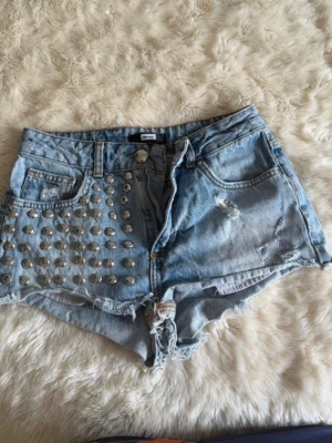 Ljusa jeansshorts med nitar - Säljer ett par ljusblå jeansshorts med hög midja, slitna detaljer och råa kanter. Ena framsidan är dekorerad med silverfärgade nitar för en edgy look. Klassisk femficksmodell i denim, perfekt till sommaren.
