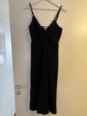 Svart klänning med knutdetalj - Stilren svart klänning med smala axelband och snygg knutdetalj framtill. Klänningen har en elegant v-ringning och är tillverkad i ett mjukt, lätt material som faller fint längs kroppen. Perfekt för dig som gillar minimalistisk och chic stil.