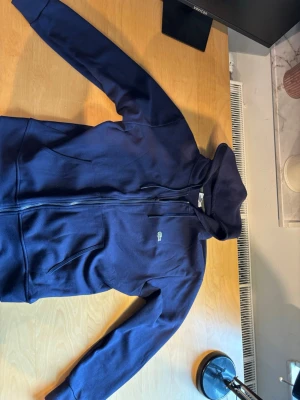 Lacoste hoodie  - Mörkblå hoodie med dragkedja framtill och två fickor. Klassisk modell med huva och små broderade detaljer på bröstet. Tillverkad i mjukt bomullsmaterial som känns skönt mot huden. Perfekt för en avslappnad och stilren look.
