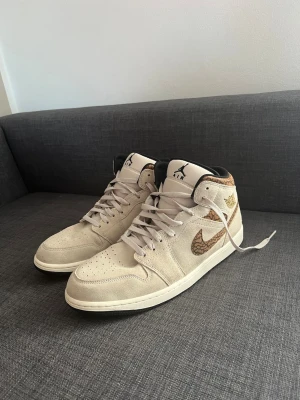 Beige Nike Air Jordan 1 sneakers - Säljer ett par beige Nike Air Jordan 1 sneakers med snygga bruna detaljer och mönstrad swoosh. Skorna har högt skaft, klassisk snörning och är tillverkade i mocka. Perfekta för dig som vill ha en unik och stilren look.